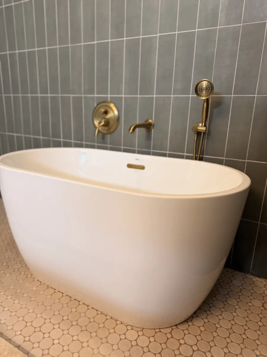 Freestanding soaking tub with brushed gold fixtures, gray stacked tile, and pebble stone floor